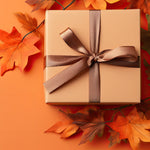 A square gift box wrapped in beige paper and tied with a wide taupe ribbon, placed on an orange background with scattered autumn leaves around it.