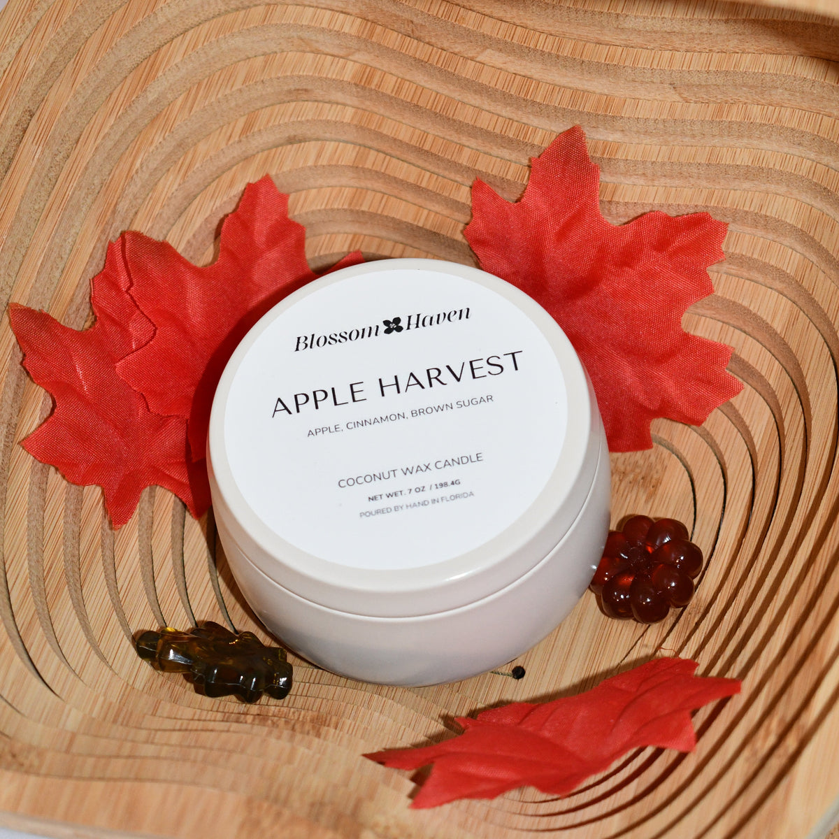 A white jar of Blossom Haven Apple Harvest coconut wax candle sits on a carved wooden tray, surrounded by red maple leaves and small amber resin pieces.
