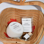 A cozy gift set from Blossom & Haven arranged in a carved wooden bowl. The set includes a white Cozy Sweater jar with a red ribbon, a wax melt package, and a small bottle of essential oil on a light gray background.