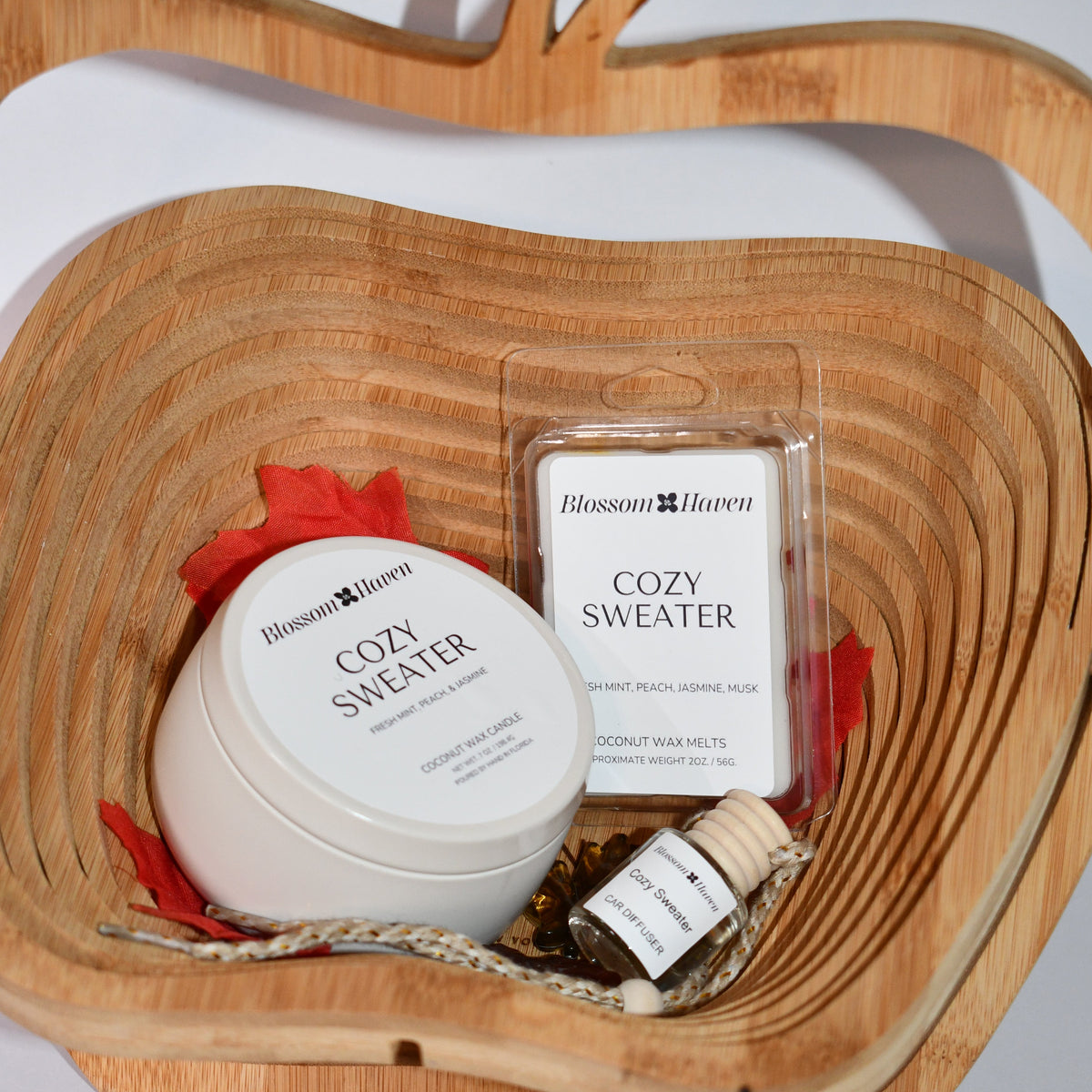 A cozy gift set from Blossom & Haven arranged in a carved wooden bowl. The set includes a white Cozy Sweater jar with a red ribbon, a wax melt package, and a small bottle of essential oil on a light gray background.