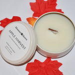 An open round metal candle tin showing pale cream wax with a small wick, next to its lid labeled 'Blossom Haven Apple Harvest,' styled with red and orange autumn leaves on a white background.