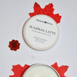 An open round candle tin showing white wax and a wick, placed beside its matching lid on a white background. The lid label reads 'Blossom Haven PUMPKIN LATTE.