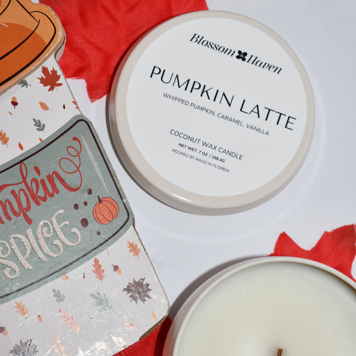 A round white candle tin labeled Blossom Haven Pumpkin Latte sits on a white surface with red autumn leaves and part of a decorative tin candle package visible at left.