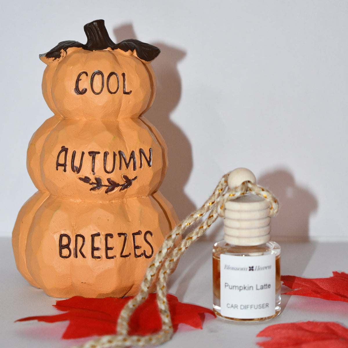 A decorative stacked pumpkin sign painted orange with the words 'COOL AUTUMN BREEZES' in black, displayed next to a small Johnson & Johnson 'Pumpkin Latte' car diffuser bottle on red paper leaves against a plain white background.