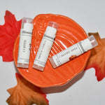 Three lip balm tubes from Blossom Haven arranged on an orange pumpkin-shaped dish with red and orange autumn leaves on a white background. The lip balms are in flavors: Coffee, Pumpkin Vanilla, and Caramel Apple.