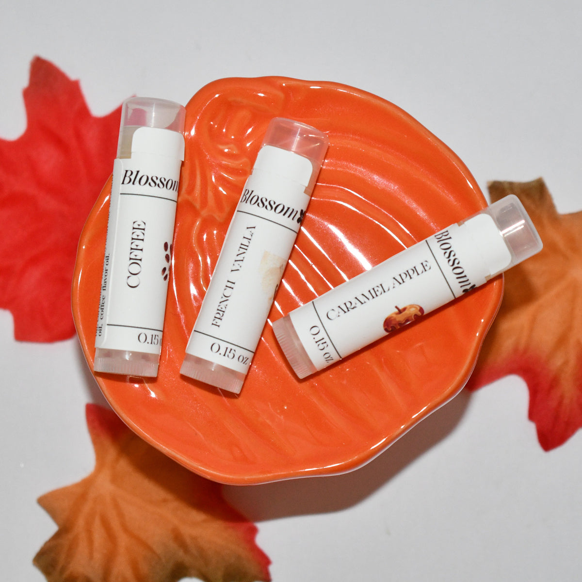 Three lip balm tubes from Blossom Haven arranged on an orange pumpkin-shaped dish with red and orange autumn leaves on a white background. The lip balms are in flavors: Coffee, Pumpkin Vanilla, and Caramel Apple.