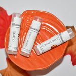 Three lip balm tubes from the brand Blossom, in Coffee, French Vanilla, and Caramel Apple flavors, arranged on an orange leaf-shaped dish with red and orange autumn leaves on a white background.
