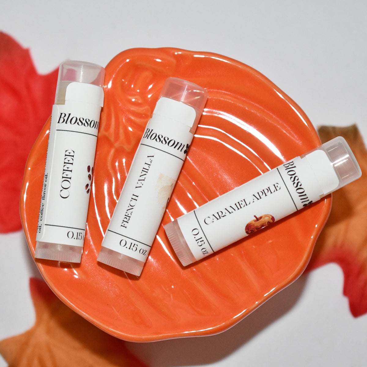 Three lip balm tubes from the brand Blossom, in Coffee, French Vanilla, and Caramel Apple flavors, arranged on an orange leaf-shaped dish with red and orange autumn leaves on a white background.
