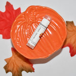 A  lip balm tube labeled 'Caramel Apple' rests on a decorative orange pumpkin-shaped dish, surrounded by red and orange autumn leaves on a white background.