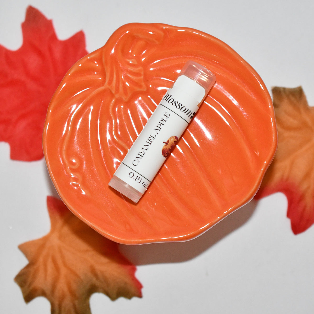 A  lip balm tube labeled 'Caramel Apple' rests on a decorative orange pumpkin-shaped dish, surrounded by red and orange autumn leaves on a white background.