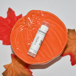 A single lip balm tube labeled 'Coffee' rests on a decorative orange pumpkin-shaped dish, surrounded by red and orange autumn leaves on a plain white background.