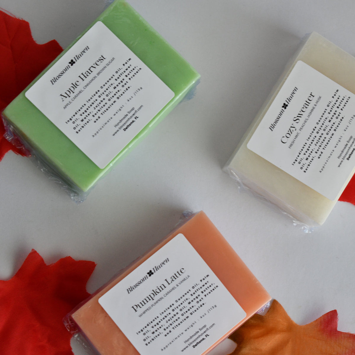 Three packaged bars of soap from arranged on a light gray surface with red and orange maple leaf cutouts. The soaps are labeled Apple Harvest (light green), Coconut Lavender (off-white), and Pumpkin Spice (orange).