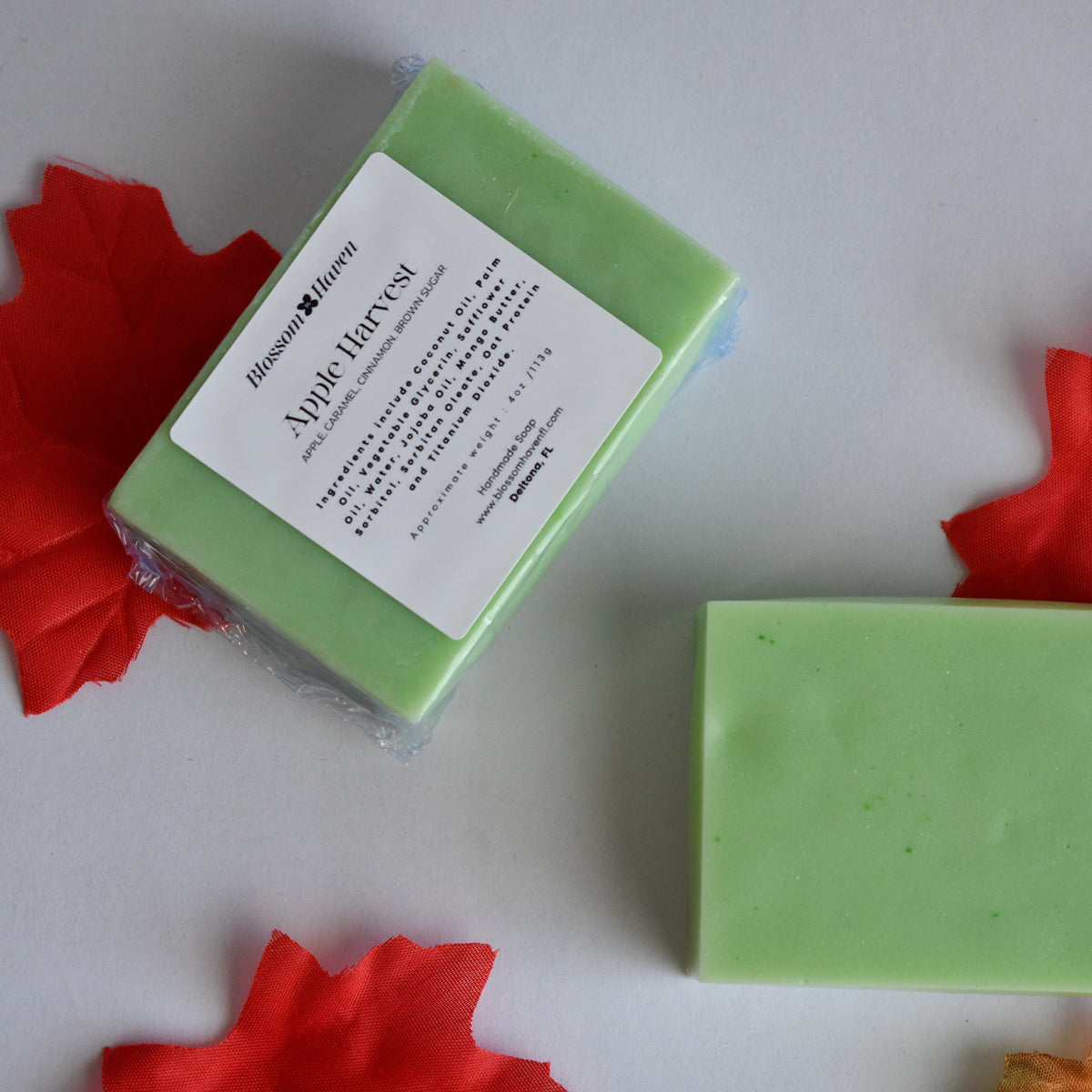 A light green rectangular bar of soap placed on a white surface next to its wrapped package, styled with red and orange autumn maple leaves around it. The label reads 'Blossom Haven' and 'Apple Harvest'.