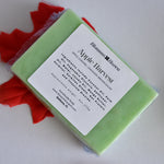 A rectangular bar of light green soap wrapped in clear plastic with a white label reading 'Blossom Haven' and 'Apple Harvest' placed on a white surface with red fabric accents.