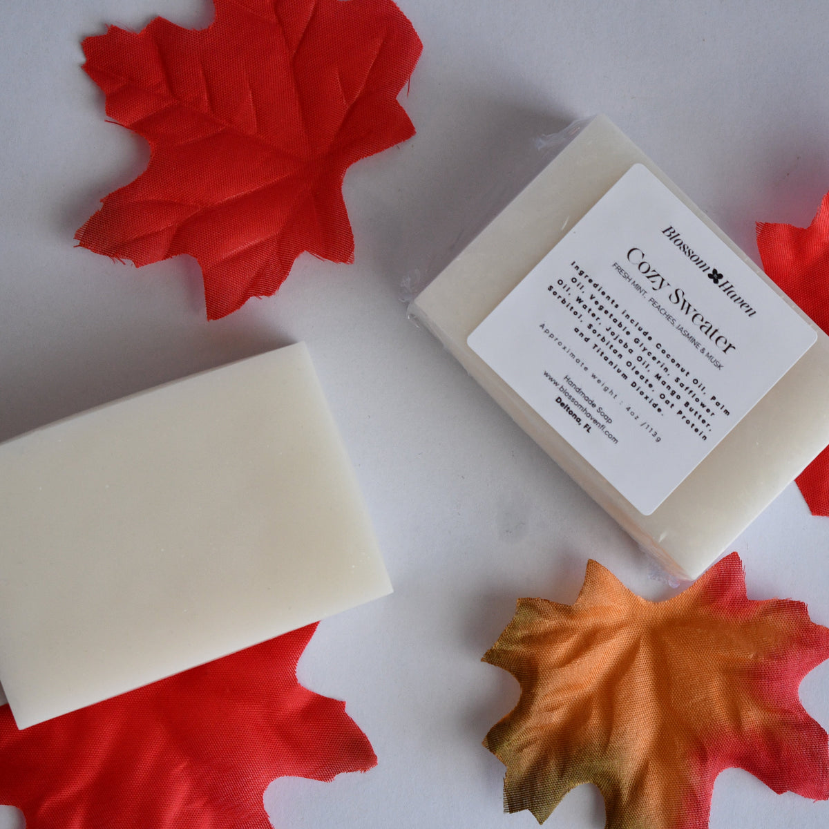 Two rectangular bars of cream-colored soap on a white surface, one unwrapped and one in its clear plastic packaging with a white label. The soap is styled with red and orange autumn maple leaves.
