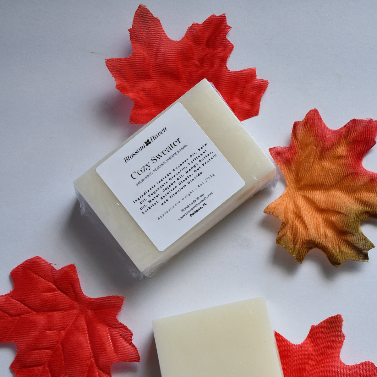 Two rectangular bars of cream-colored soap arranged on a light gray surface with decorative autumn leaves. One bar is plain, and the other is wrapped in clear plastic with a white label reading 'Cozy Sweater'.