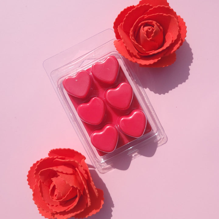 Red heart-shaped wax melt in a clear plastic container with two red roses on a pink background