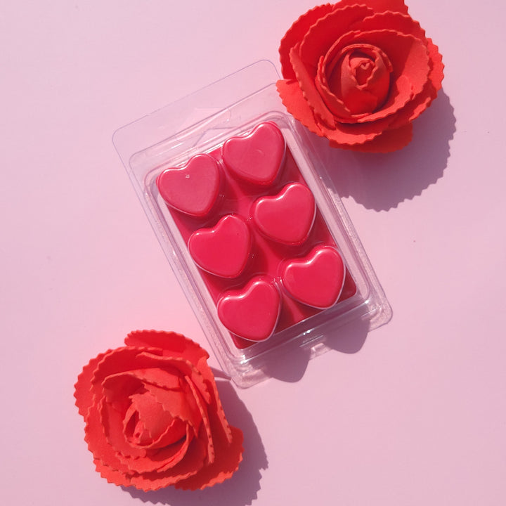 Pack of pink heart-shaped wax melts with two red roses on a pink background.